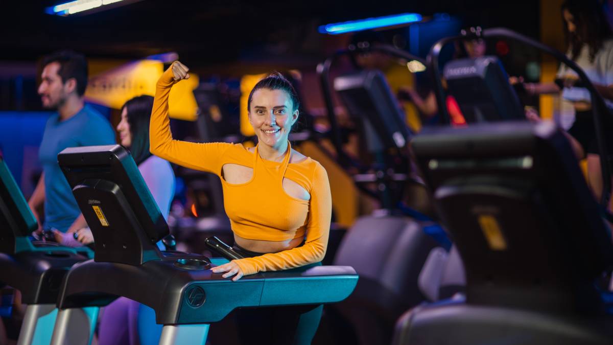 mujer parada sobre una caminadora de un gimnasio Smart Fit con un brazo recargado en el aparato y el otro flexionado hacia arriba enseñando su bicep en señal de fuerza