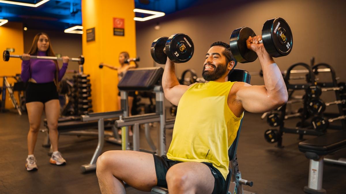 hombre realizando un press militar con dos mancuernas, sentado en un banco dentro del area de peso libre de un gimnasio smart fit