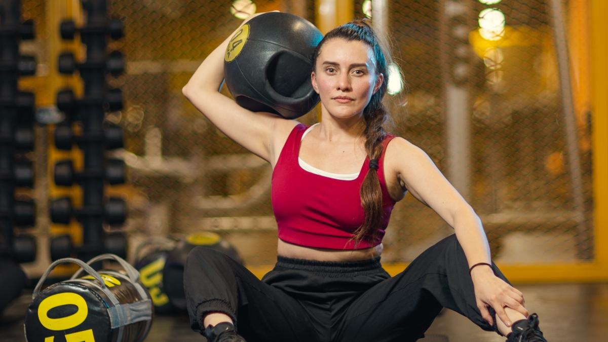 mujer sentada en el suelo de un gimnasio smart fit con un balón medicinal apoyado en su hombro y sostenido con una mano, la otra mano en us pierna