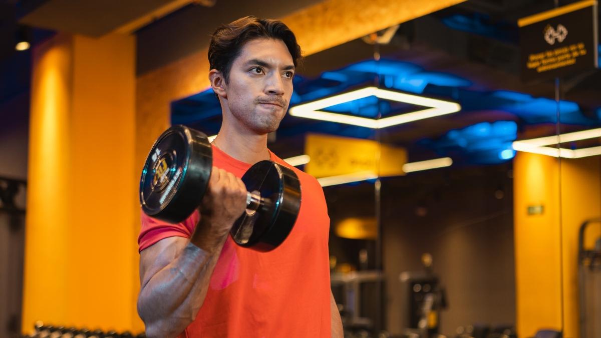 Hombre joven levantando una mancuerna en un gimnasio, concentrado en su entrenamiento de fuerza. Lleva una camiseta roja y esta rodeado de un ambiente moderno con iluminacion azul y amarilla. El fondo muestra maquinas y equipos de gimnasio, creando un entorno motivador para el ejercicio