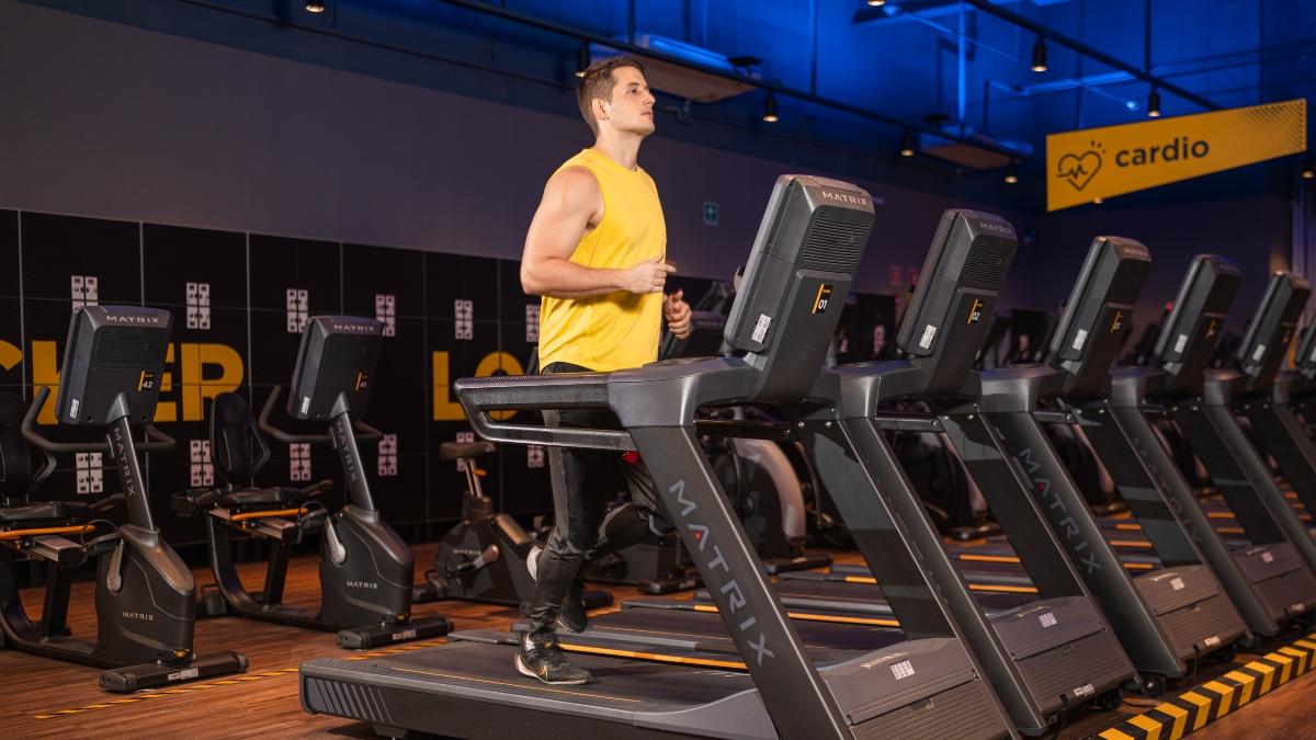 Hombre joven con camiseta sin mangas amarilla corriendo en una caminadora en un area de cardio en el gimnasio Smart Fit, rodeado de varias maquinas de ejercicio en un ambiente moderno