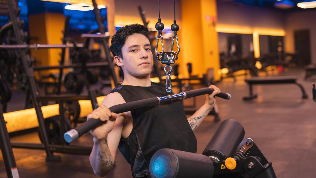 Un hombre joven en camiseta sin mangas realiza un ejercicio de lat pulldown en una maquina en un gimnasio Smart Fit, rodeado de equipo de pesas.