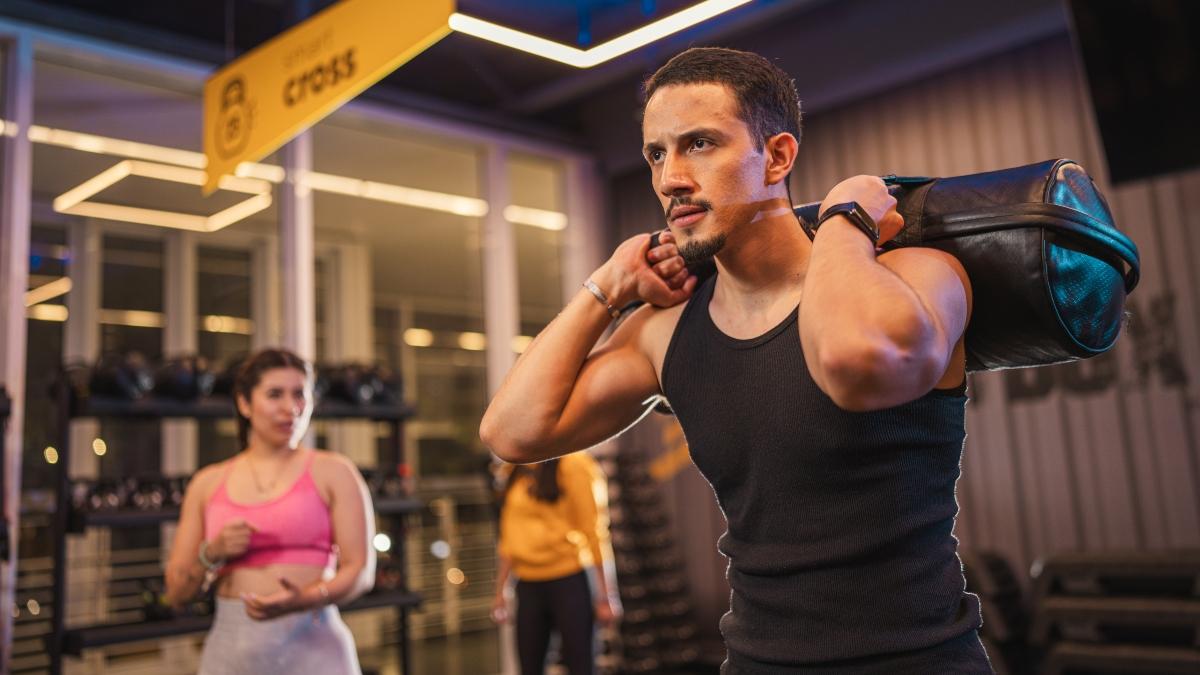Hombre joven con camiseta sin mangas negra sosteniendo un saco de entrenamiento sobre sus hombros en el gimnasio Smart Fit. Al fondo, una mujer en camiseta rosa observa mientras realiza su propio ejercicio