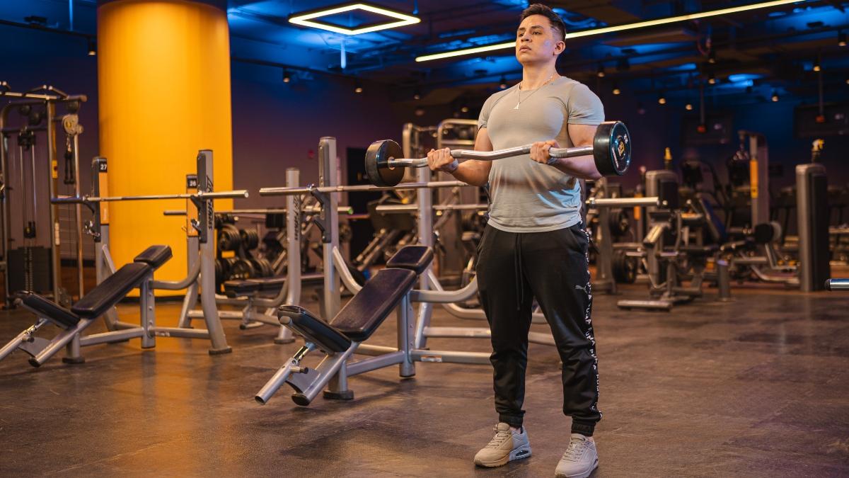 Hombre realizando ejercicios isotonicos de curl de biceps de pie con barra en un gimnasio Smart Fit, rodeado de equipo moderno en un ambiente amplio y bien iluminado.