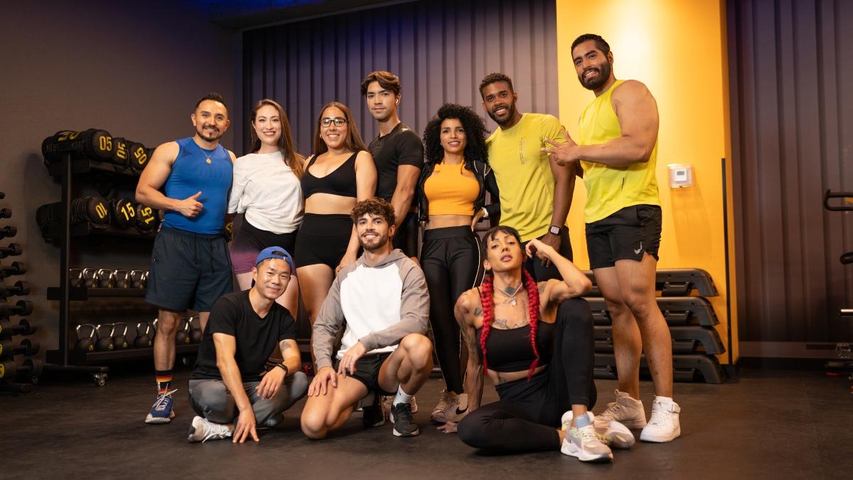 Grupo diverso de personas sonrientes posando en un gimnasio moderno con equipo de entrenamiento en el fondo, representando el concepto de comunidad activa en el mercado fitness en Mexico y gimnasios Smart Fit