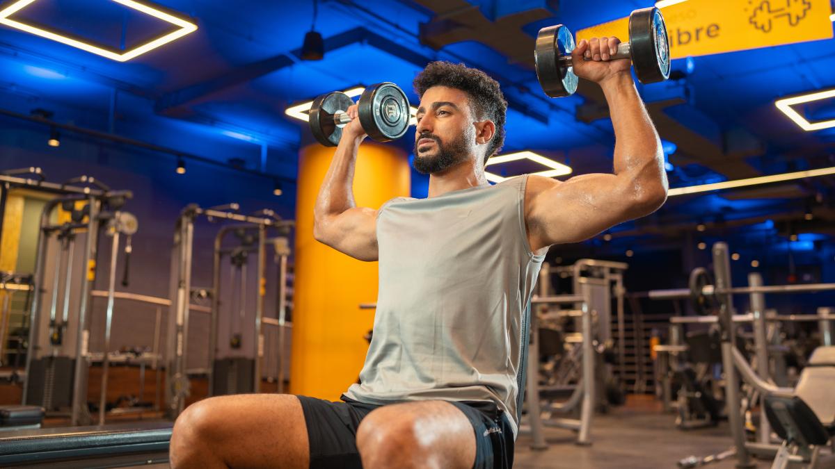 Hombre realizando press militar con mancuernas en un gimnasio Smart Fit, demostrando la tecnica de drop set en un entorno moderno y bien iluminado.